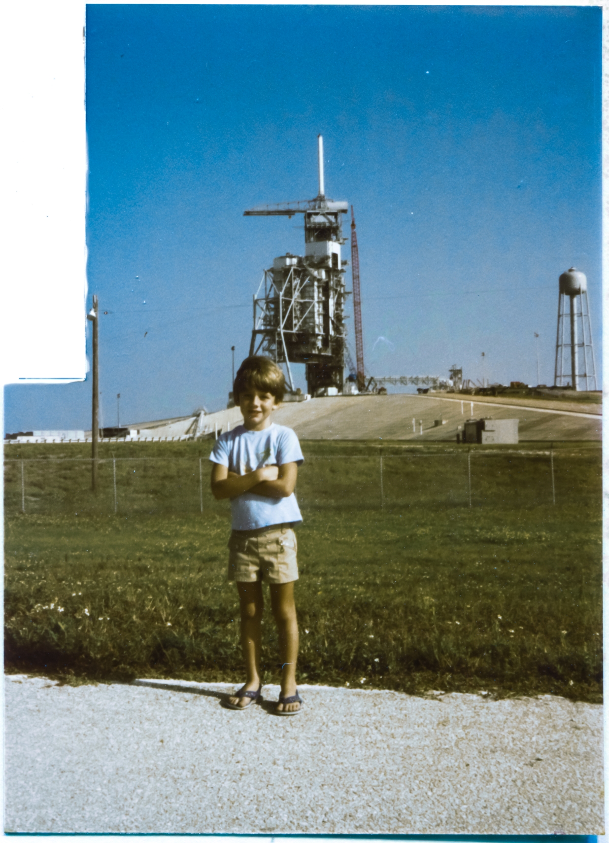 Image 073. At an age of 6 years old, an age that could not be more perfectly matched for the moment, Kai MacLaren stands on the Bypass Road just outside the Perimeter Fence of Space Shuttle Launch Complex 39-B, Kennedy Space Center, Florida. Behind him, to the northeast, the looming structures of the RSS and the FSS tower above the concrete body of the Pad, attended by large construction cranes. Kai is as much in his element as it is ever possible to be, and understands exactly how rare and sublime a moment this is, and will remember it for a lifetime. As will his father. Photo by James MacLaren.